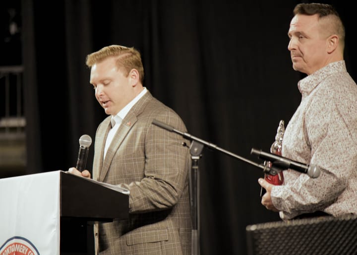 A speaker in a plaid blazer at the lectern beside a guest holding an award