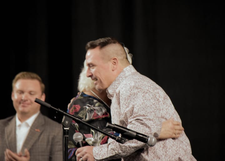Two people embracing on stage while a nearby guest applauds