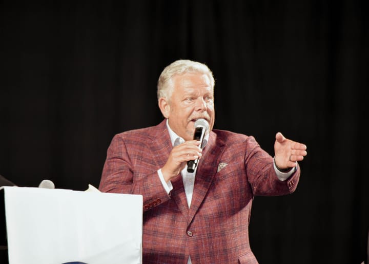 A speaker in a burgundy plaid blazer extending a hand toward the audience